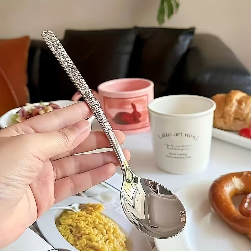 Hand holding a silver spoon with a blurred background of a table with food and cups.