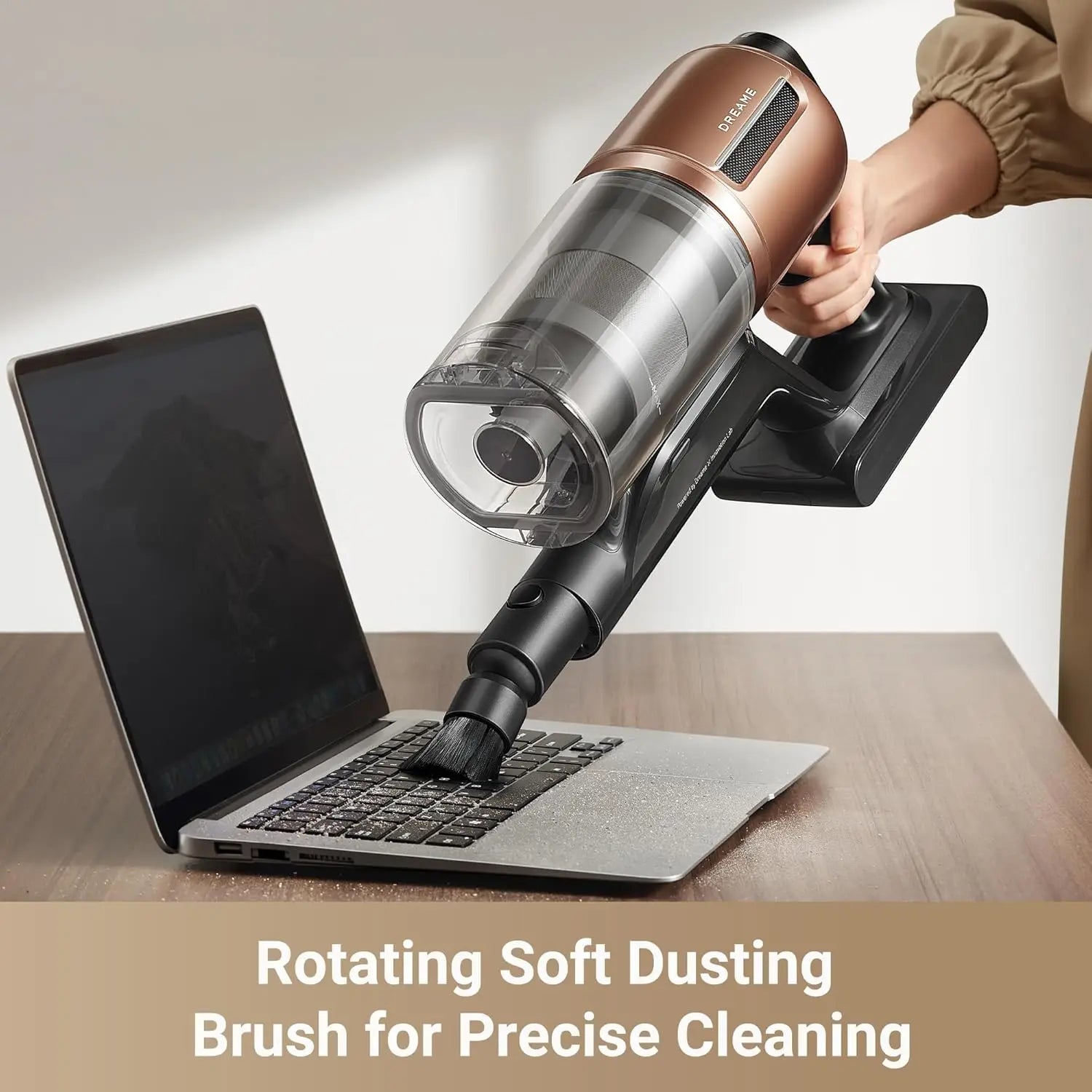 Person using a handheld vacuum cleaner with a rotating soft dusting brush to clean a laptop on a wooden desk.