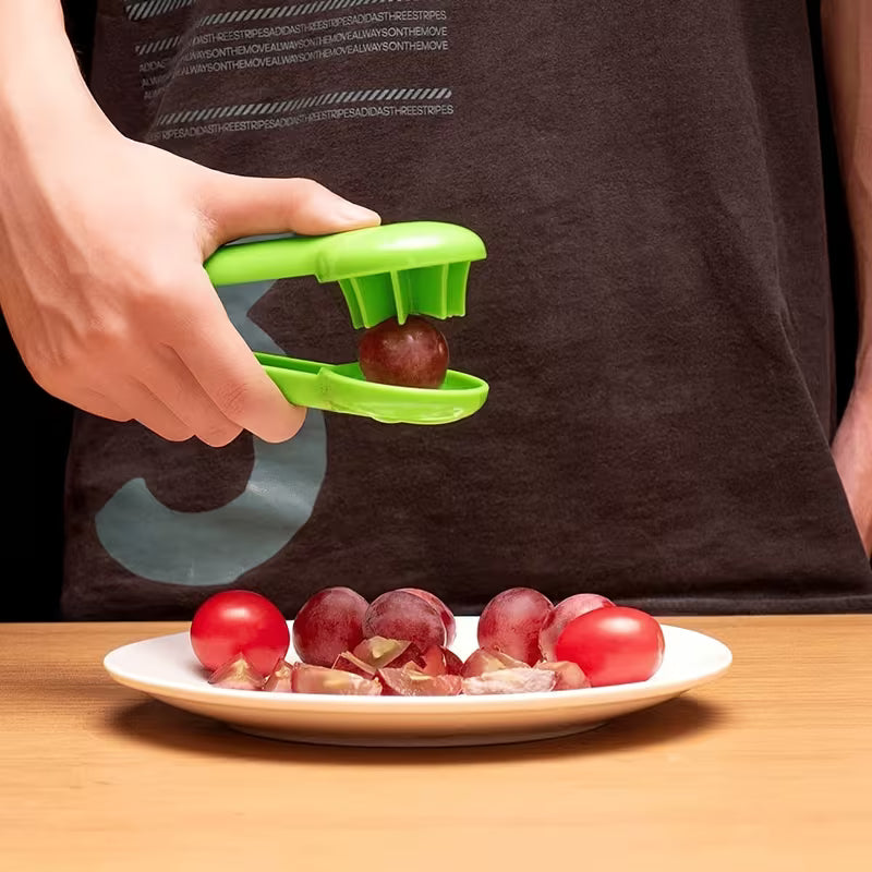 Person using a green grape peeler on red grapes with a plate of peeled grapes and tomatoes in the background.