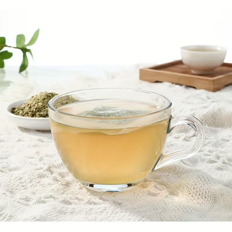 Clear glass cup filled with yellow tea on a textured surface with green leaves and a wooden tray in the background.