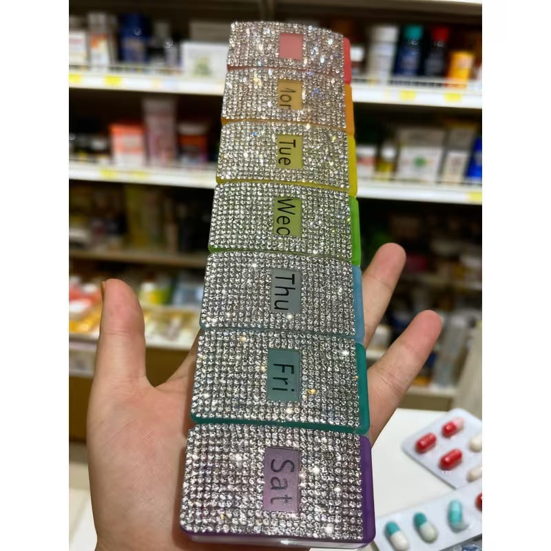 Hand holding a weekly pill organizer with rhinestone embellishments in a pharmacy setting